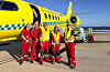 Fem personer i røde og gule redningsdrakter står foran et gult ambulansefly på en flyplass.