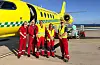 Fem personer i røde og gule redningsdrakter står foran et gult ambulansefly på en flyplass.