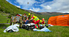 Redningspersonell i refleksvester og uniformer arbeider rundt en båre på en gresslette med fjell og telt i bakgrunnen.