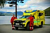 To personer i røde arbeidsklær står foran en gul Ford Ranger-ambulanse ved vannet.