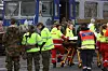 Flere personer i refleksvester og militæruniform løfter en båre foran en buss.