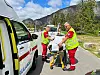 To personer i røde og gule vester triller en båre mellom en ambulanse og en transportbil utendørs.