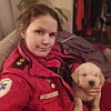 Kvinne i rød ambulanseuniform holder en liten, lys valp foran seg innendørs.