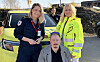 To helsearbeidere står bak en sittende mann foran en ambulanse.