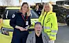 To helsearbeidere står bak en sittende mann foran en ambulanse.