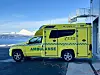 Gul og grønn UNN-ambulanse parkert ved en bygning med fjell og sjø i bakgrunnen.